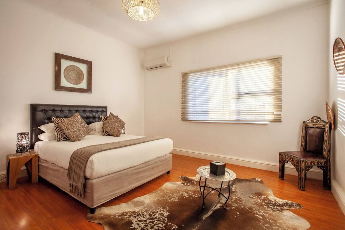 A bedroom with a large bed, a bedside table, a chair, a window with blinds, and a ceiling light fixture.