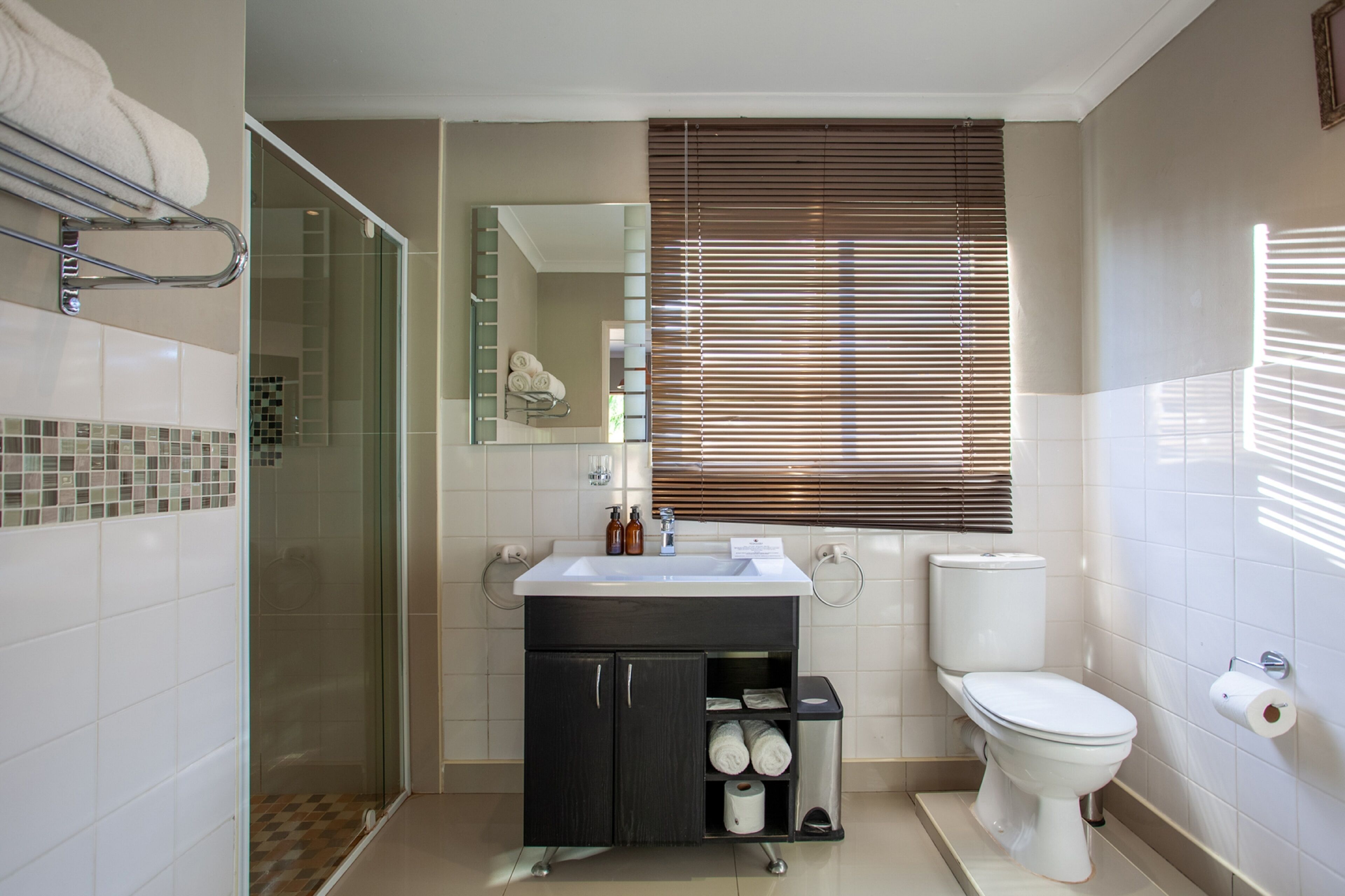 A modern bathroom with a glass shower enclosure, a sink with toiletries, a toilet, and a window with blinds.