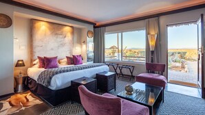 A hotel room with a large bed, a desk, a chair, and a view of the desert landscape. - Inverdoorn Private Game Reserve (Breede River)