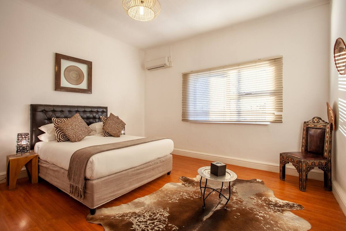 A bedroom with a large bed, a small round table, a chair, a window with blinds, and a wall-mounted air conditioner.