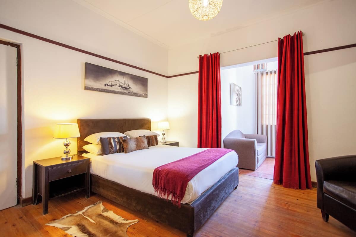 A hotel room with a large bed, two bedside lamps, a red curtain, a leather sofa, and a mounted painting.
