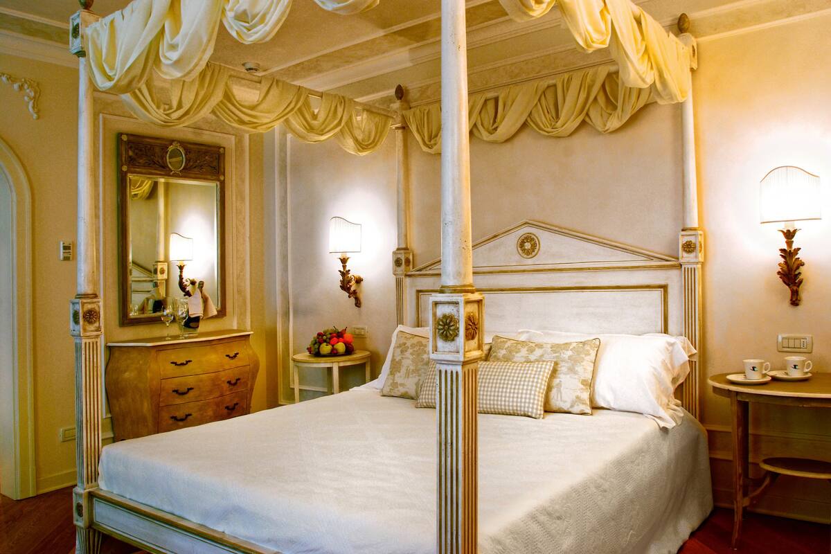 Una cama con dosel, un aparador de madera, una mesa redonda con fruta y un espejo con marco decorativo.