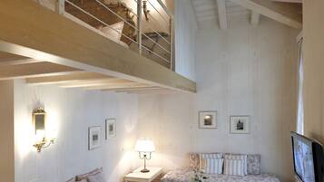 Een ruime loft-achtige kamer met een hoge plafond, houten balken, een inbouwtrappenhuis, twee banken, een salontafel en een televisie.