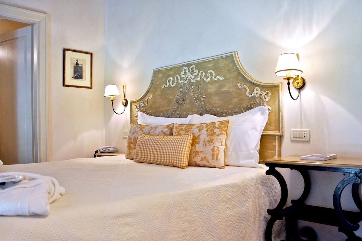 Una cama bien hecha con edredón blanco y almohadas decorativas, una mesita de noche con lámpara y un cuadro en la pared.