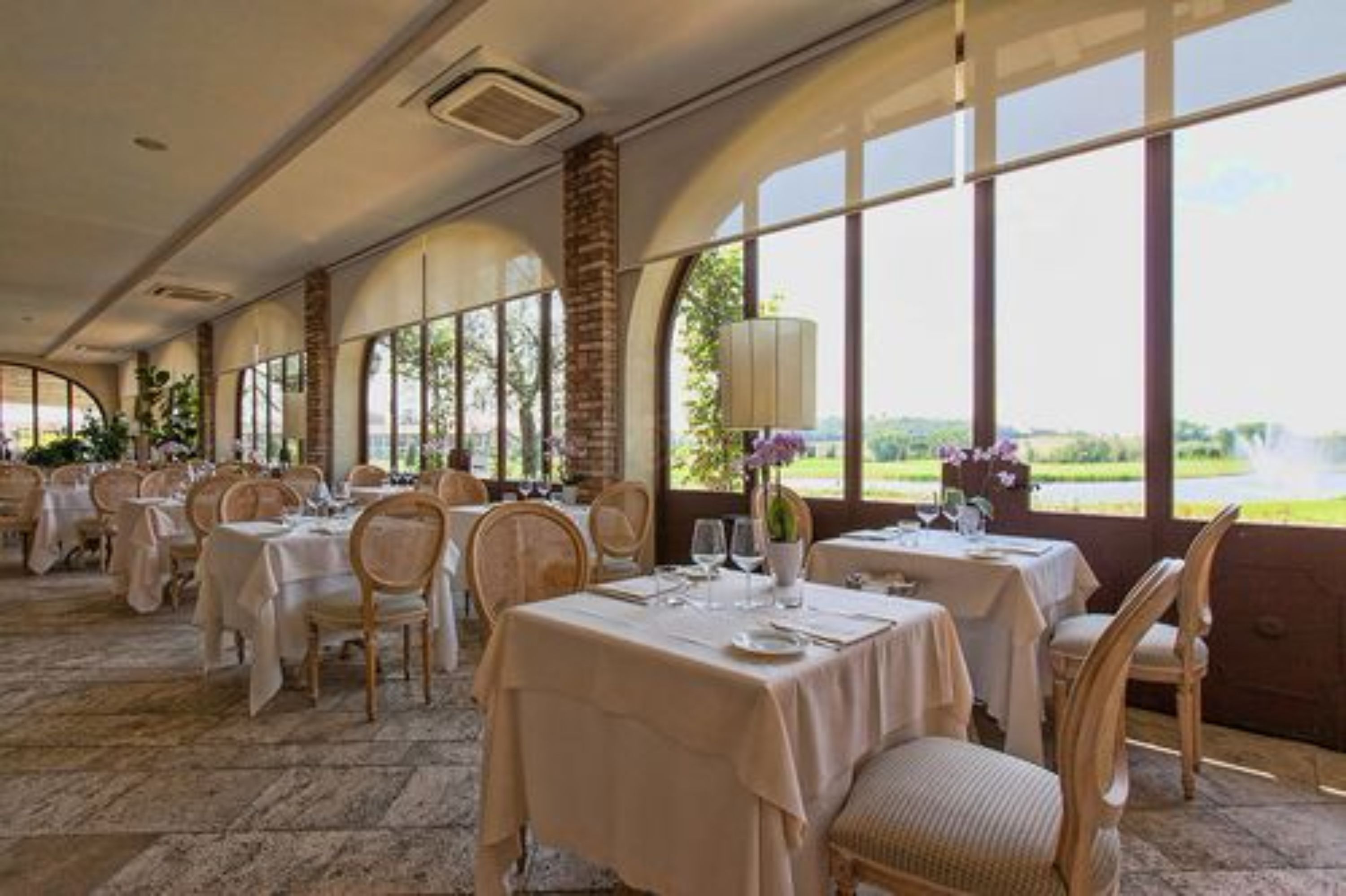 A dining area with tables set for a meal, large windows offering a view of greenery, and a brick column.