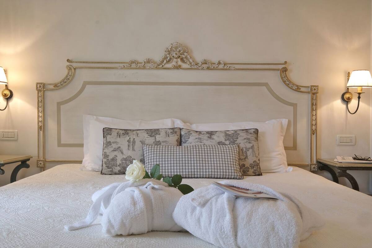 Una cama bien hecha con sábanas blancas, dos almohadas con estampados diferentes, una rosa blanca y una revista.