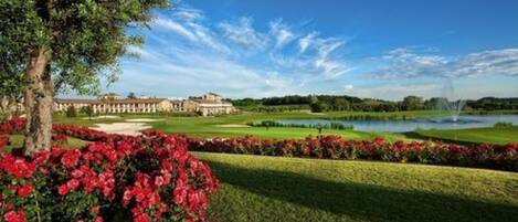 Un campo de golf con un edificio grande al fondo, rodeado de céspedes bien cuidados y una masa de agua.