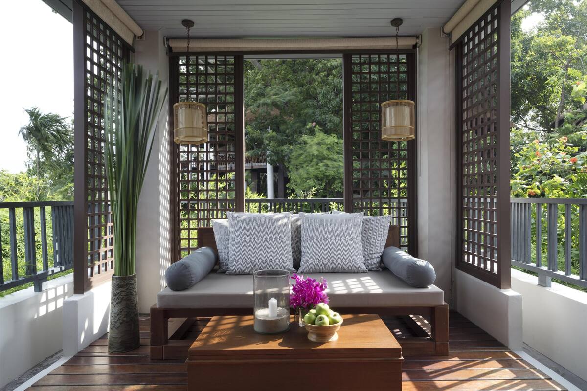 A balcony with a wooden floor, a sofa with cushions, a glass candle holder, a vase with flowers, and a bowl of fruit.