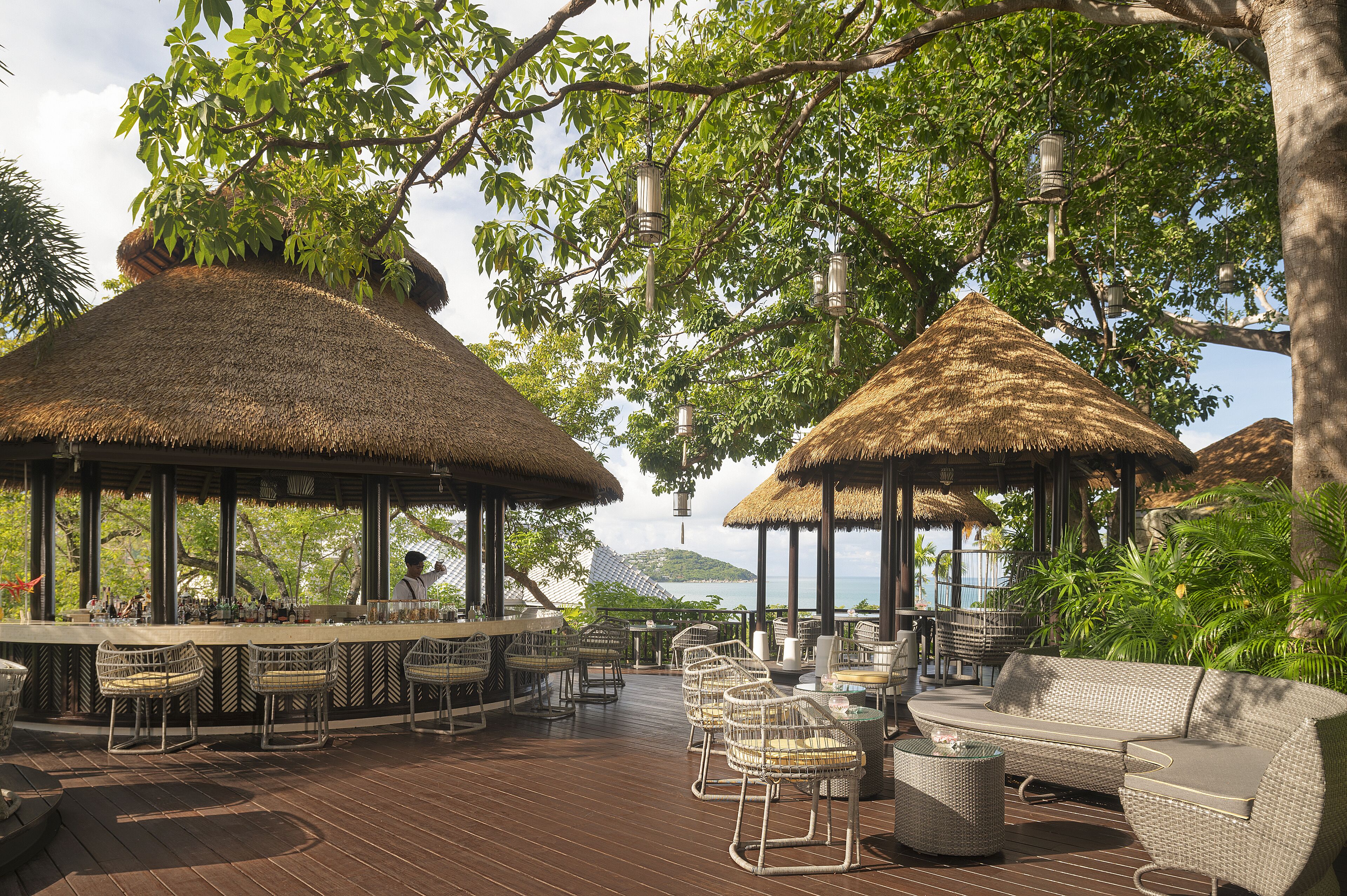 A covered outdoor bar area with thatched-roof structures, wicker furniture, and a view of the sea.