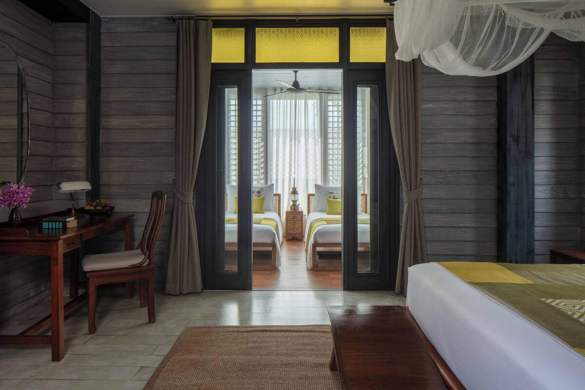 A hotel room with a bed, a desk with a chair, a small table with a lamp, and a view through a doorway to another room with a bed and a table.