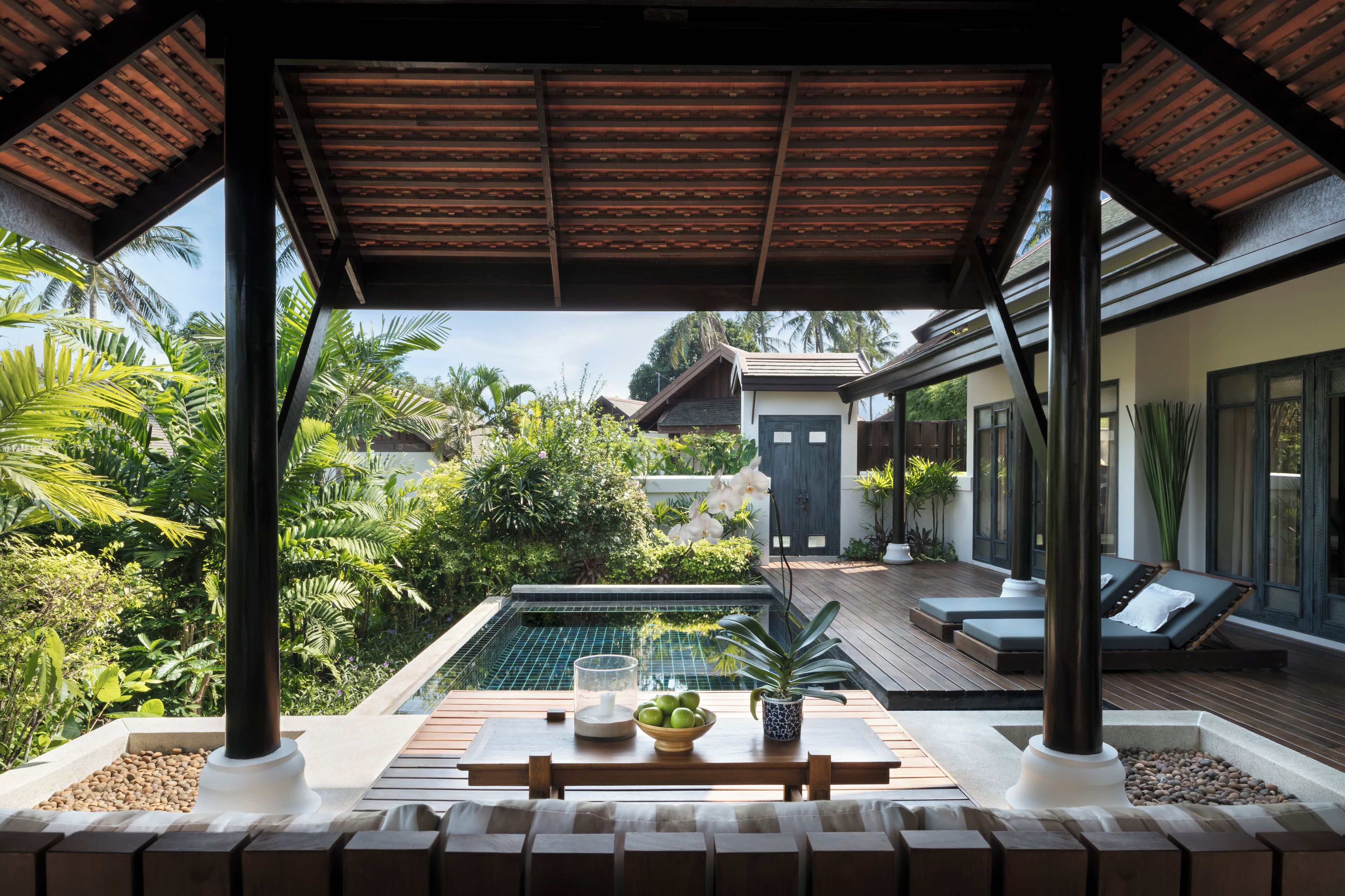 A covered outdoor area with a pool, lounge chairs, and a table with fruits and a bowl.