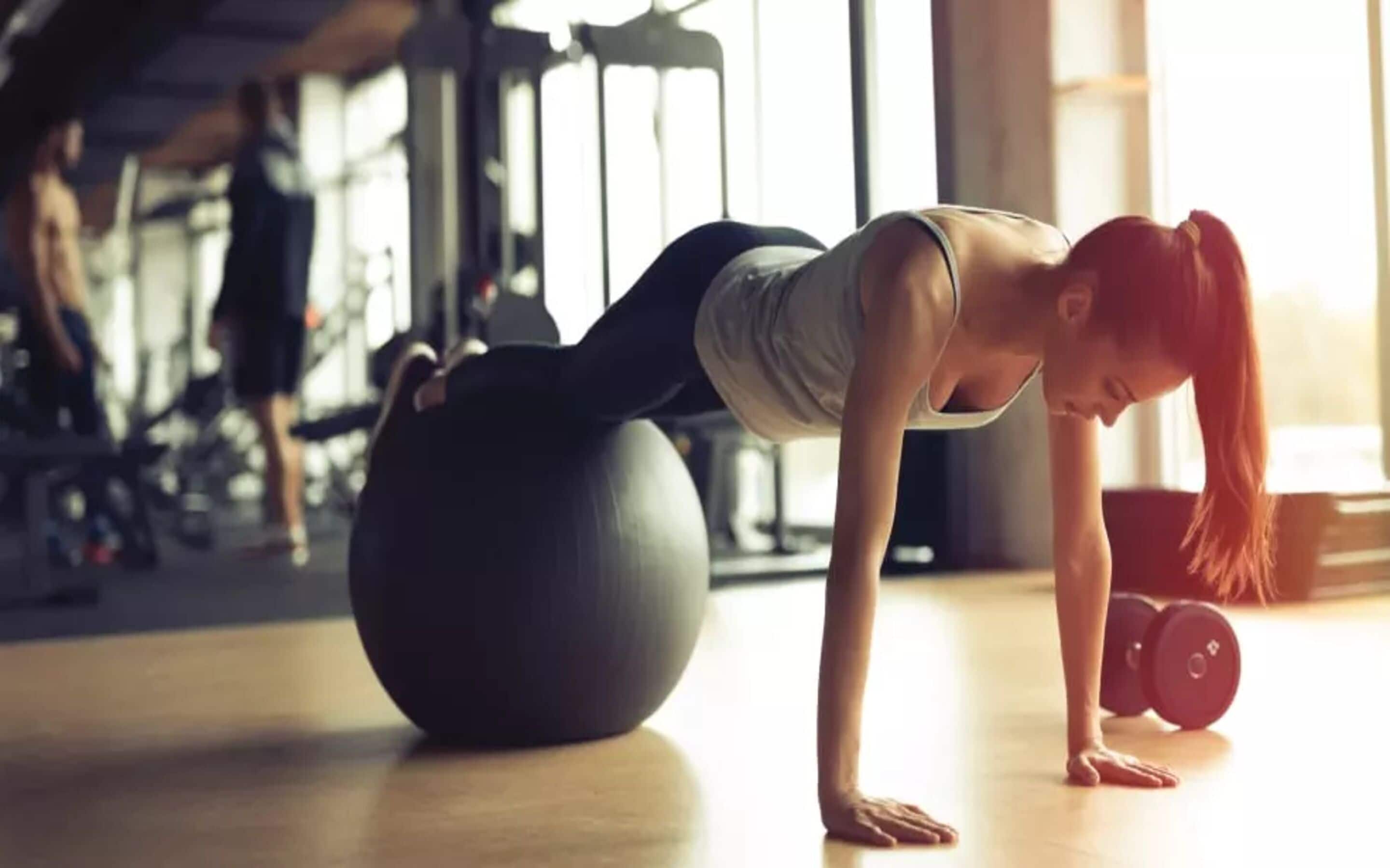 Una persona haciendo ejercicio en una pelota de gimnasia grande en un gimnasio con varios equipos al fondo.