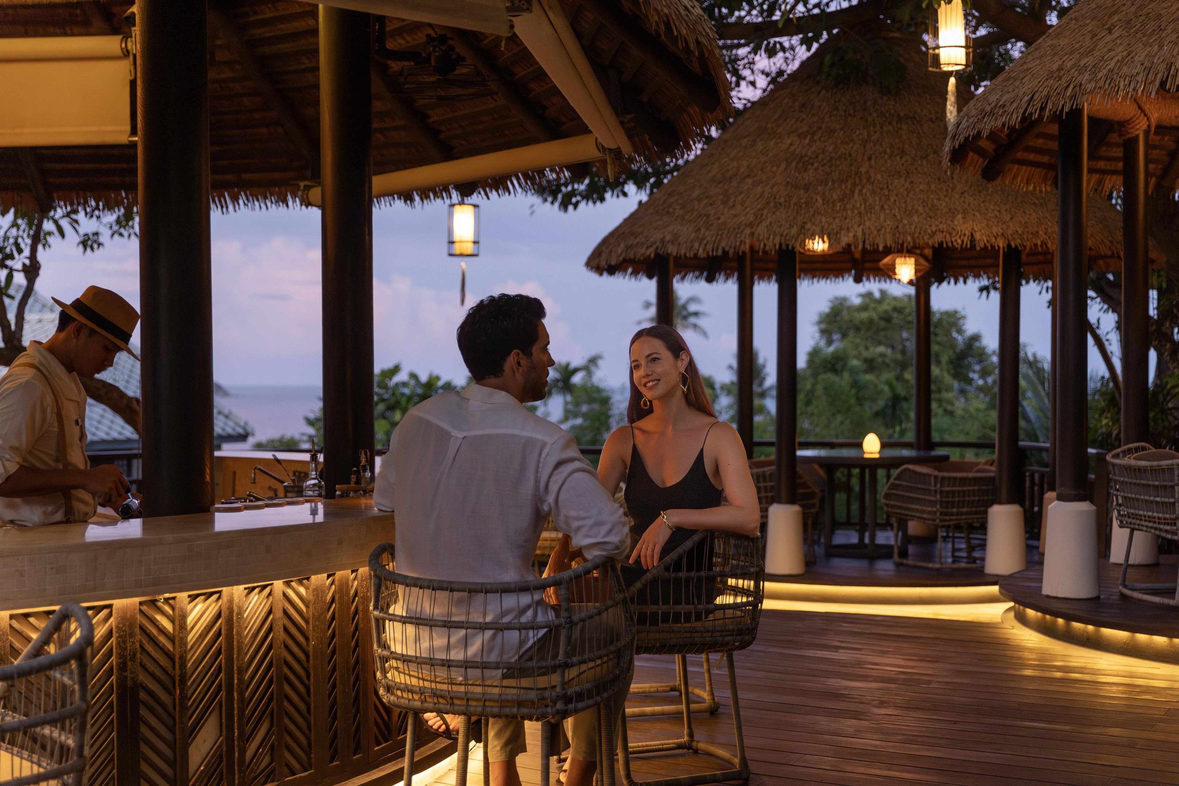 A wooden deck with thatched-roof structures, outdoor seating, and a view of the ocean.
