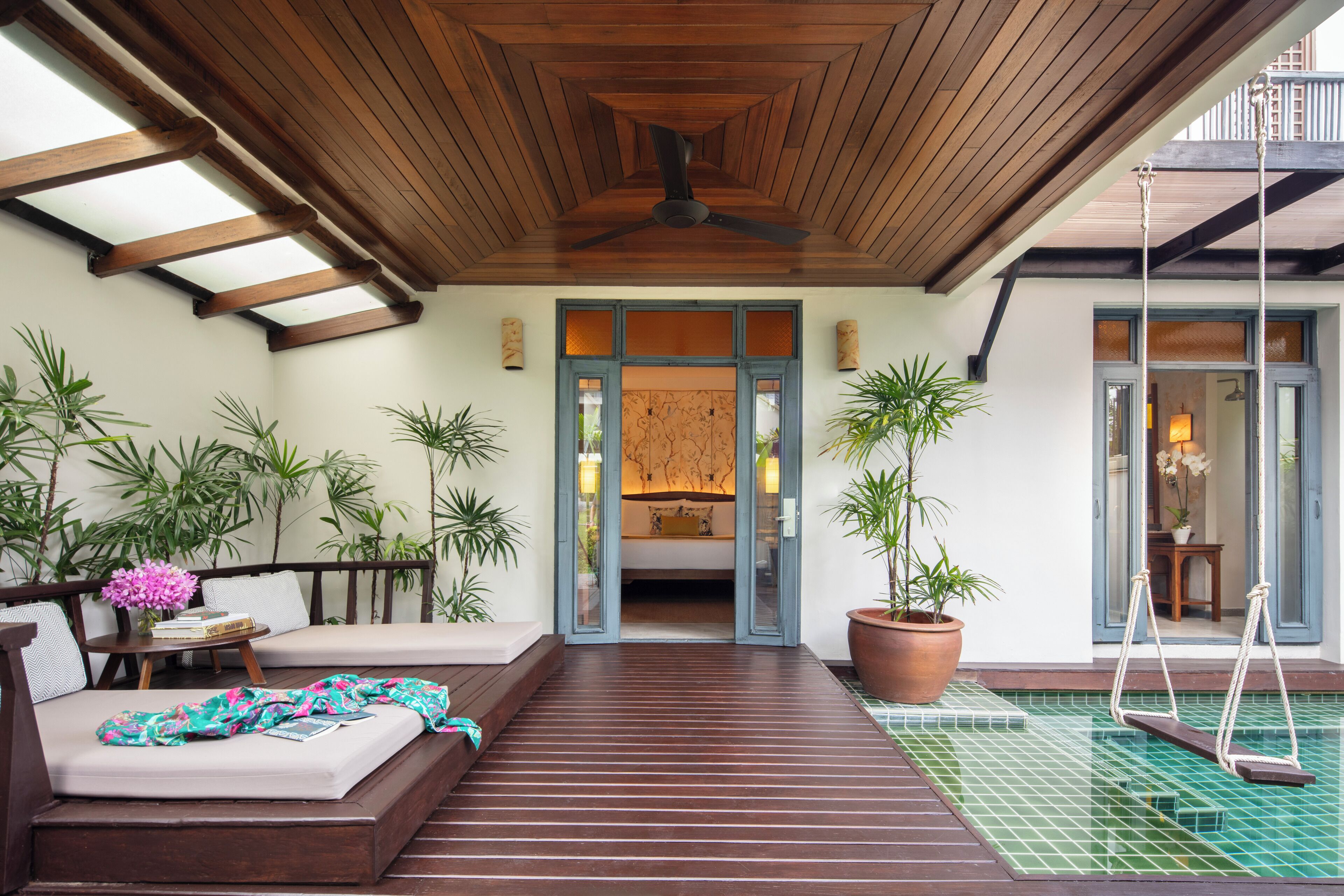A wooden deck with a ceiling fan, a sofa with cushions, a small table with a vase of flowers, and a large potted plant.