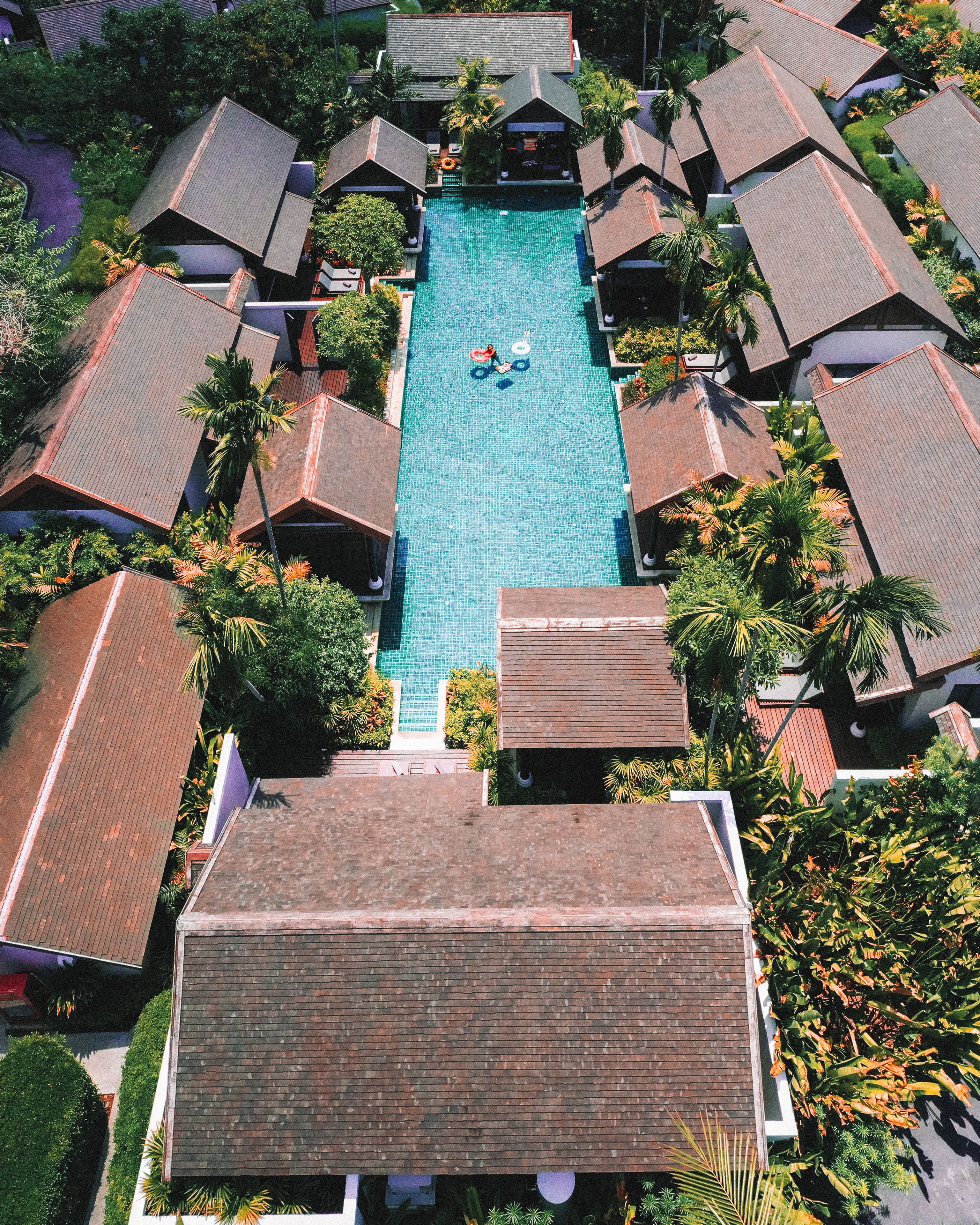 An aerial view of a resort with a central swimming pool surrounded by bungalow-style accommodations with sloped roofs.