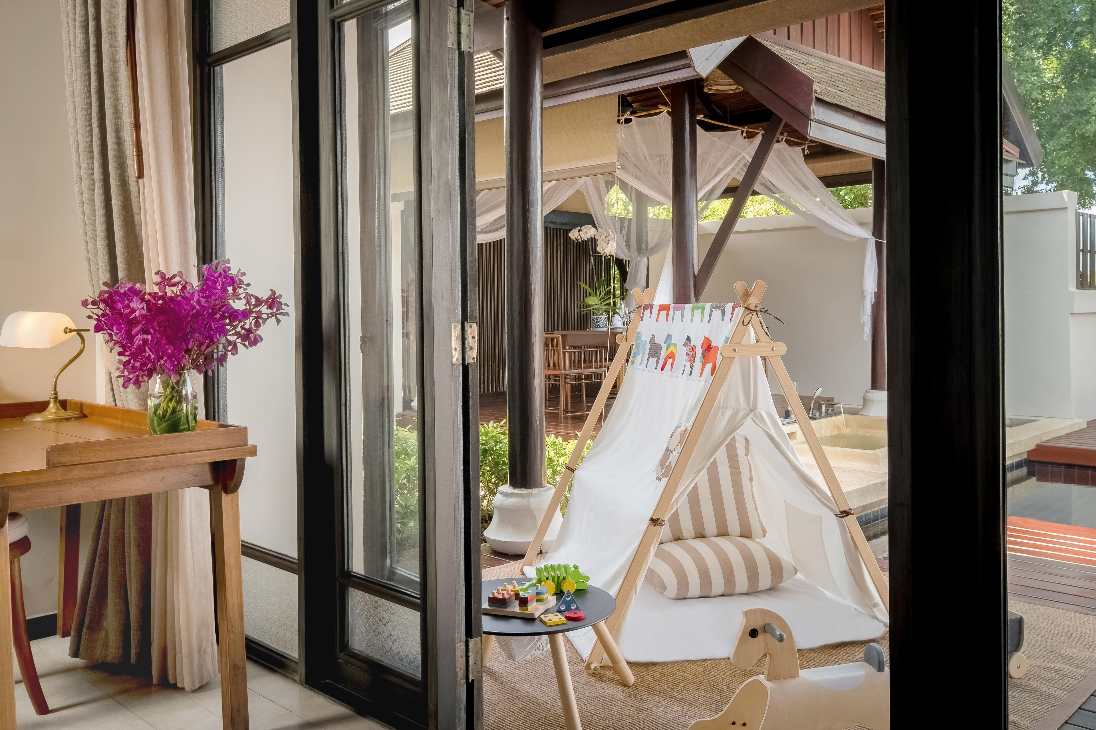 Una habitación con un ventanal grande, una mesa de madera con una lámpara, un jarrón de flores y una zona de juegos infantil con una tienda de campaña y juguetes.
