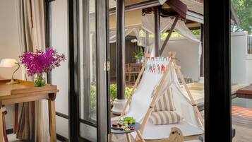 A room with a large window, a wooden table with a lamp, a vase of flowers, and a children's play area with a tent and toys.