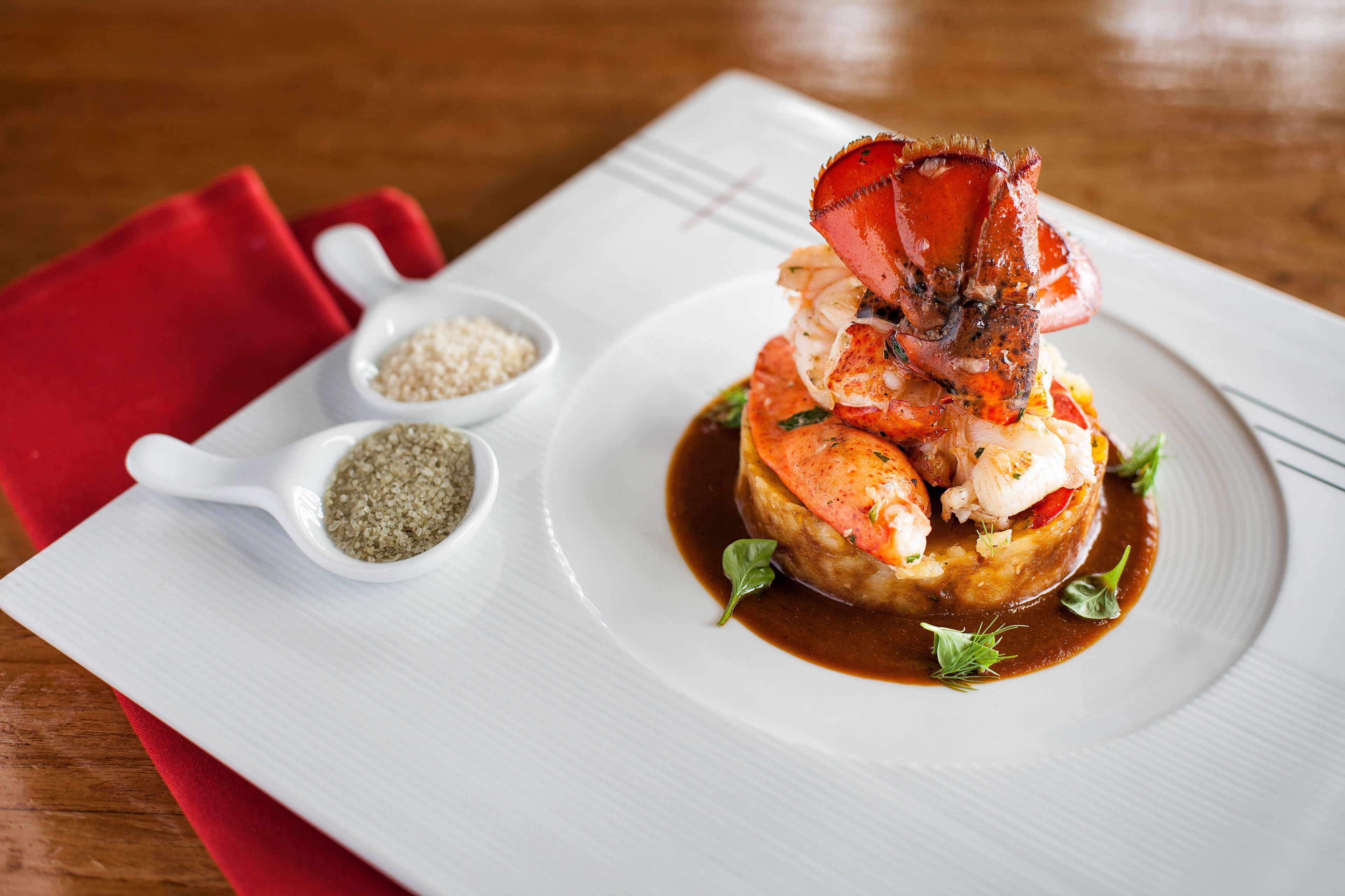 Un plat de homard servi dans une assiette blanche, accompagné de deux petits ramequins de condiments placés sur une serviette rouge à côté.