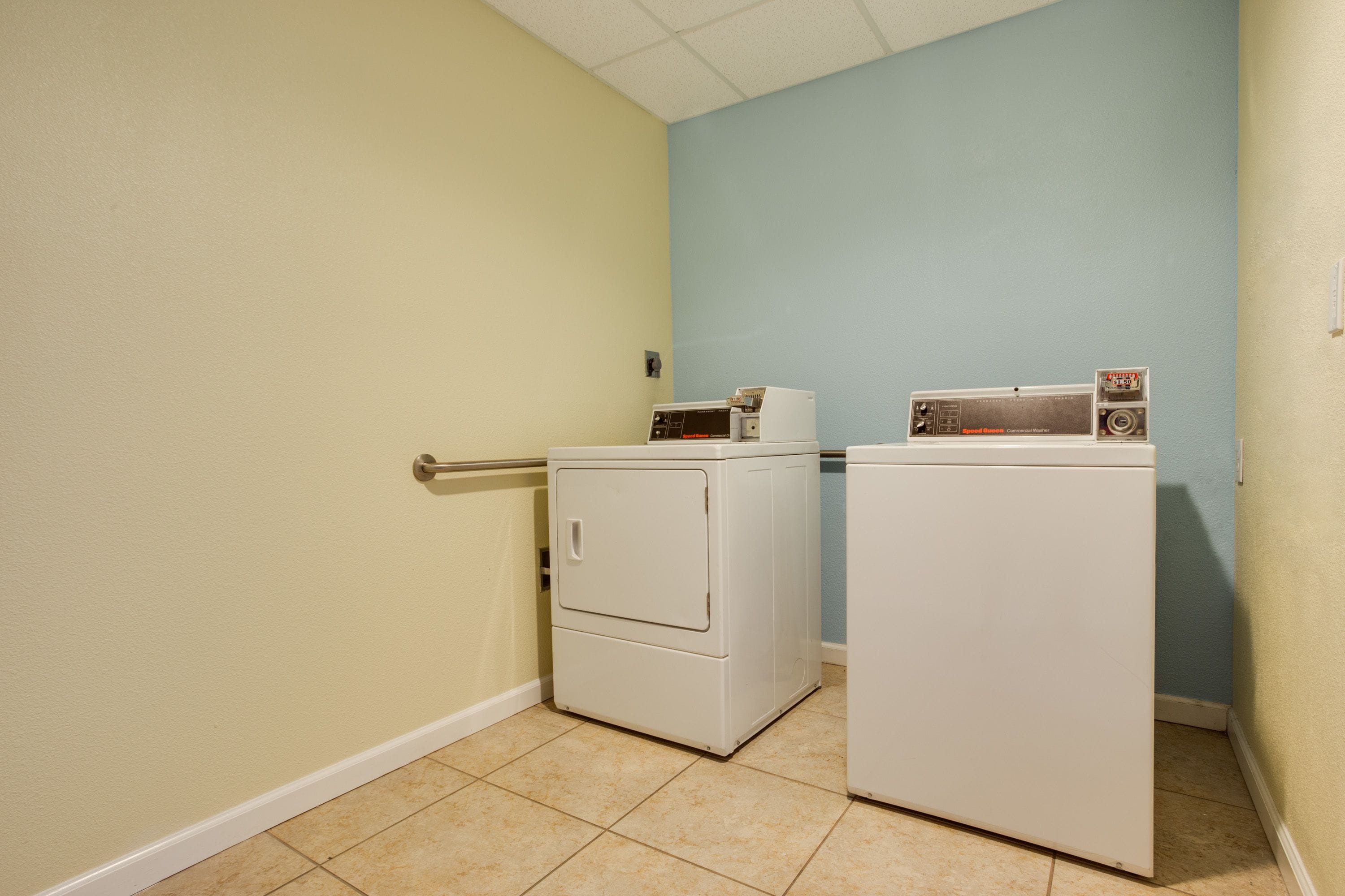 Une buanderie avec deux machines à laver et deux sèche-linge blancs, un sol carrelé de couleur beige, des murs vert clair et bleu, et une porte blanche.