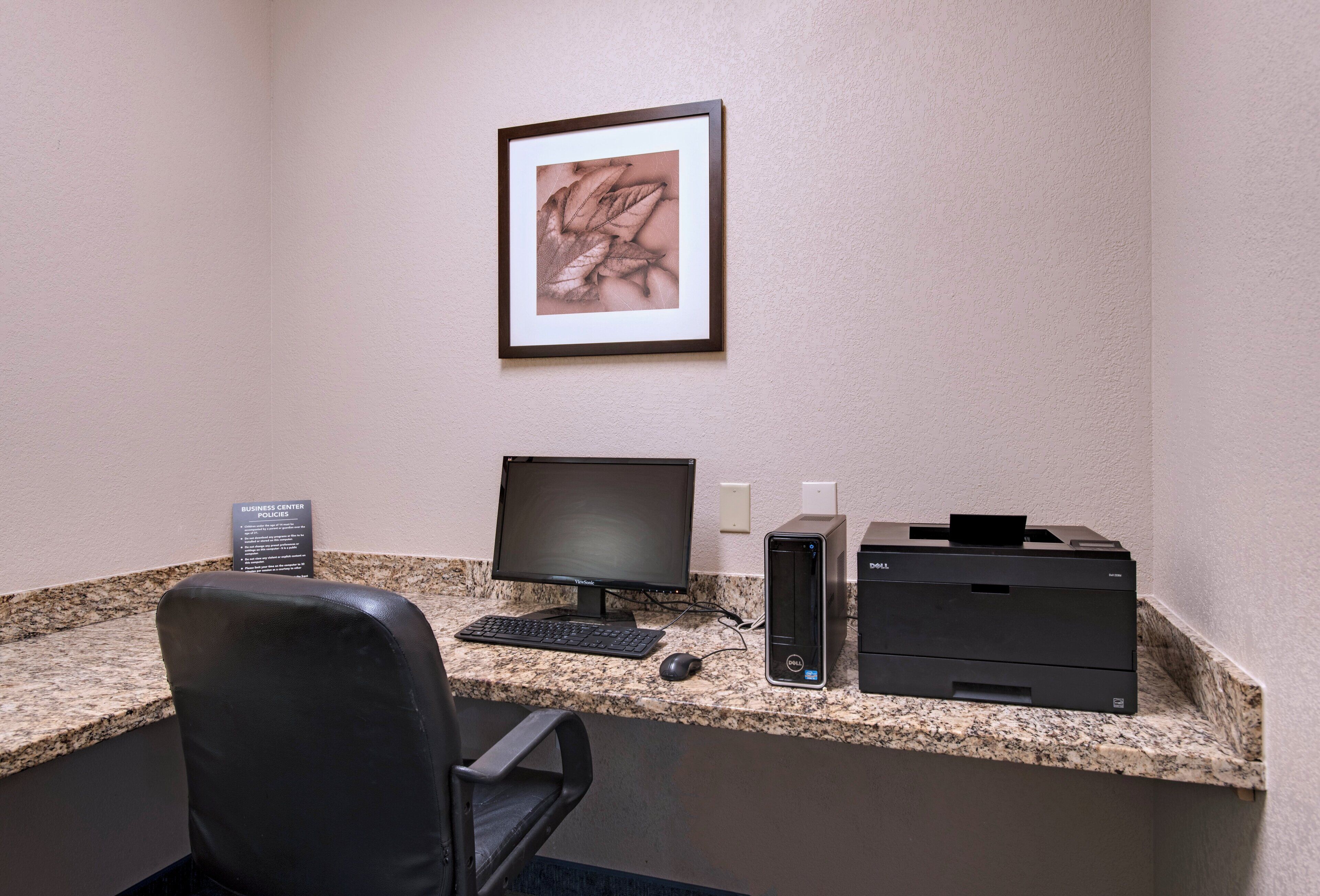 A business center with a desktop computer, a monitor, a keyboard, a printer, and a framed picture on the wall.