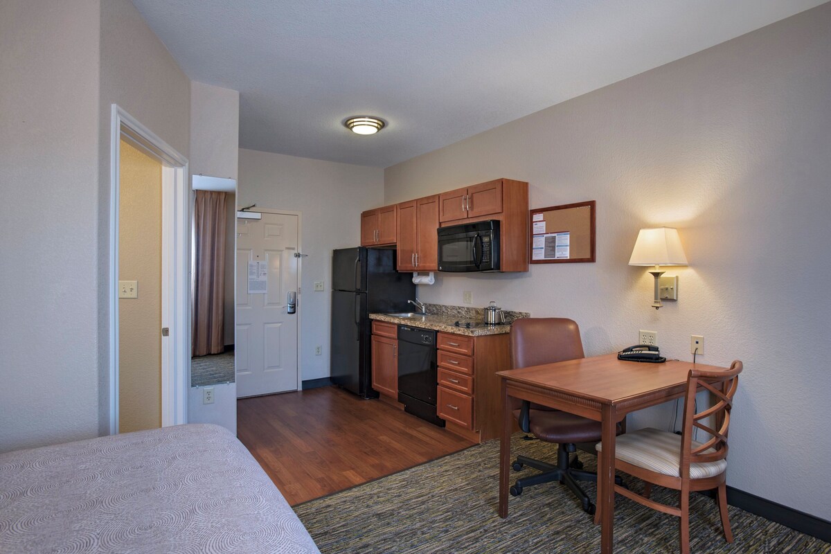 Een compacte hotelkamer met kitchenette, een eethoek met tafel en stoelen, een bed en een badkamer met douchegordijn.