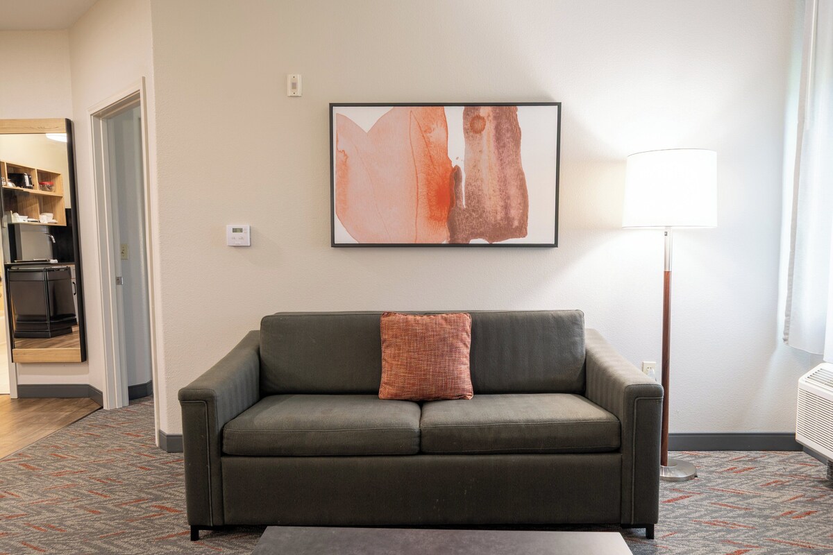 Ein Wohnzimmer mit einer Couch, einem gerahmten abstrakten Gemälde, einer Lampe und einem gemusterten Teppich.