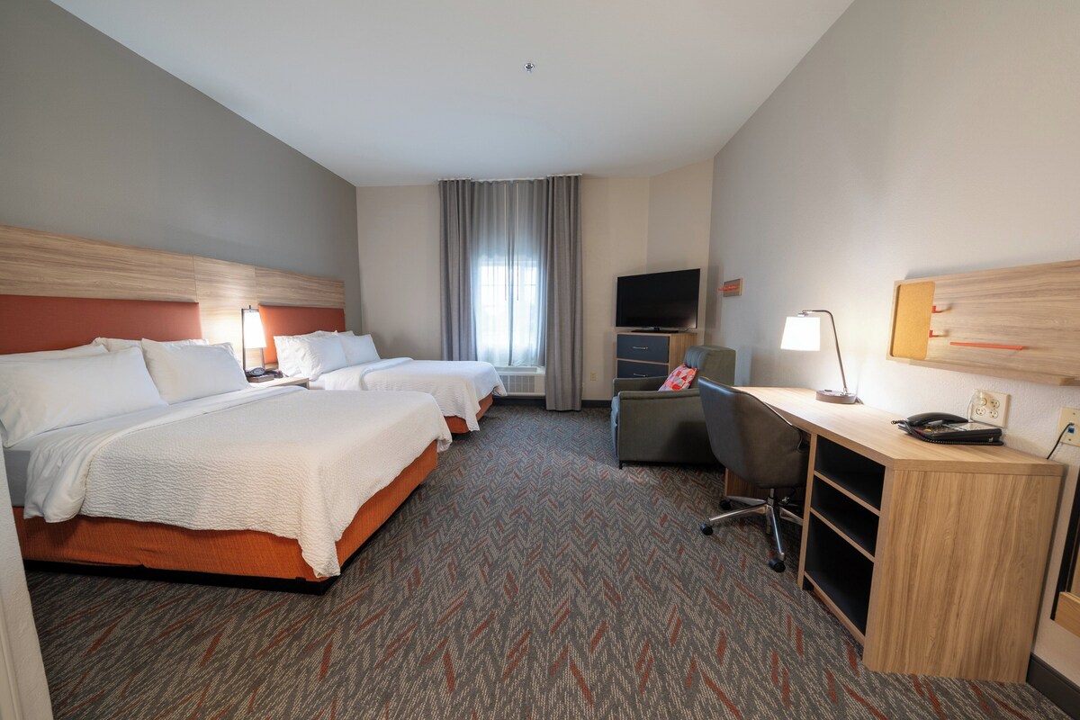 Ein Hotelzimmer mit zwei Betten, einem Schreibtisch, einem Stuhl, einem Fernseher und einem Fenster mit Vorhängen.