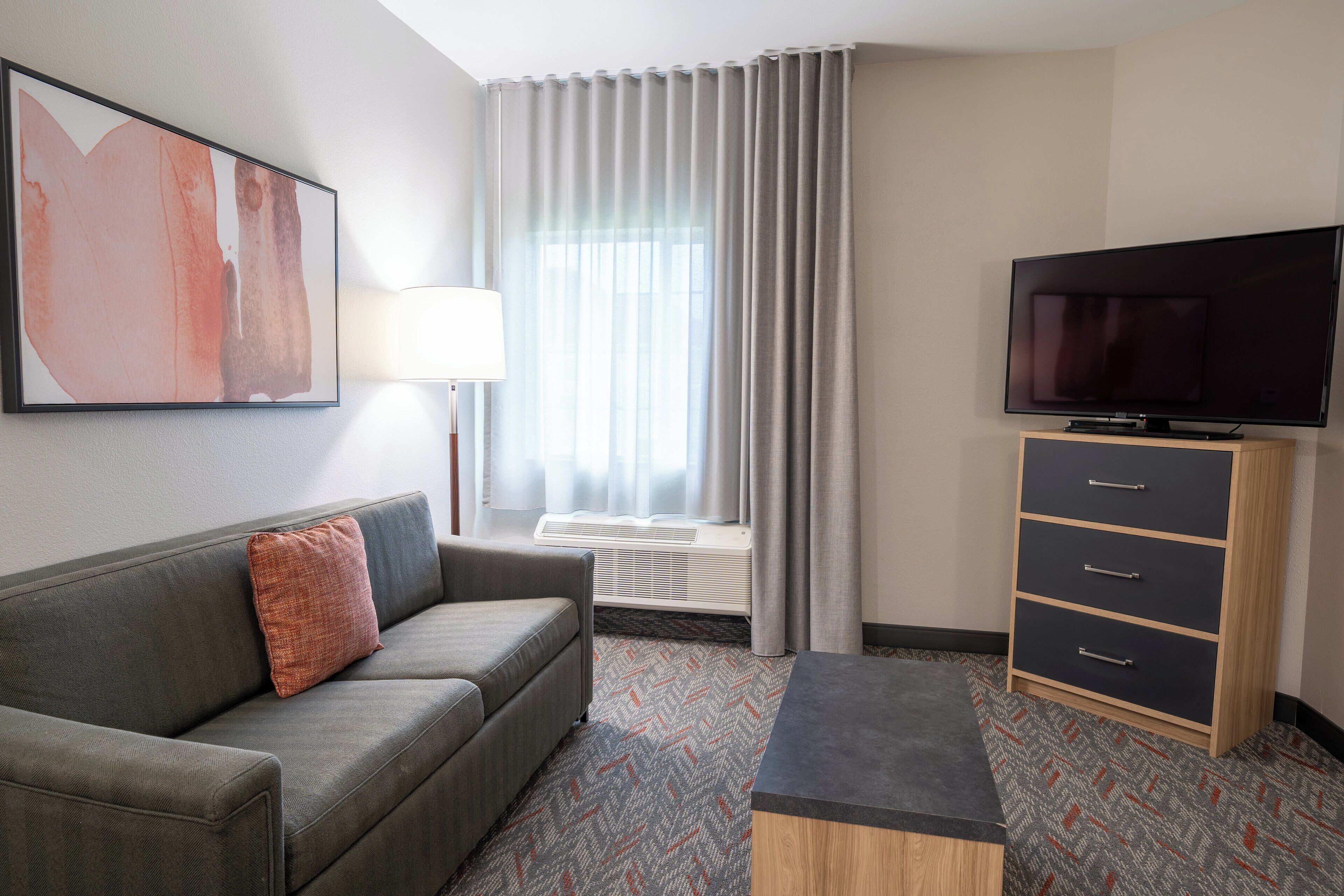 A hotel room with a gray sofa, a flat-screen TV on a wooden stand, a patterned carpet, and a large window with curtains.