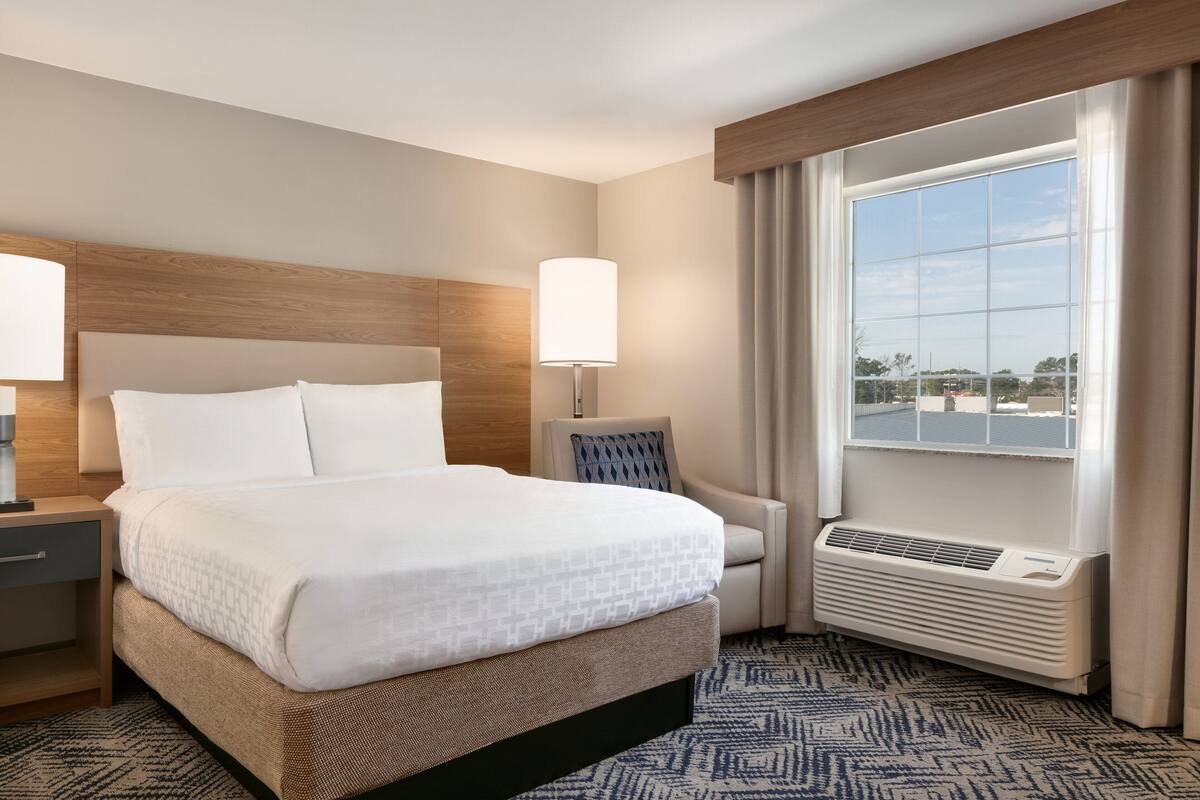Ein Hotelzimmer mit einem großen Bett, Nachttischen, einem Sessel, einem Fenster mit Vorhängen und einer Wand-Klimaanlage.