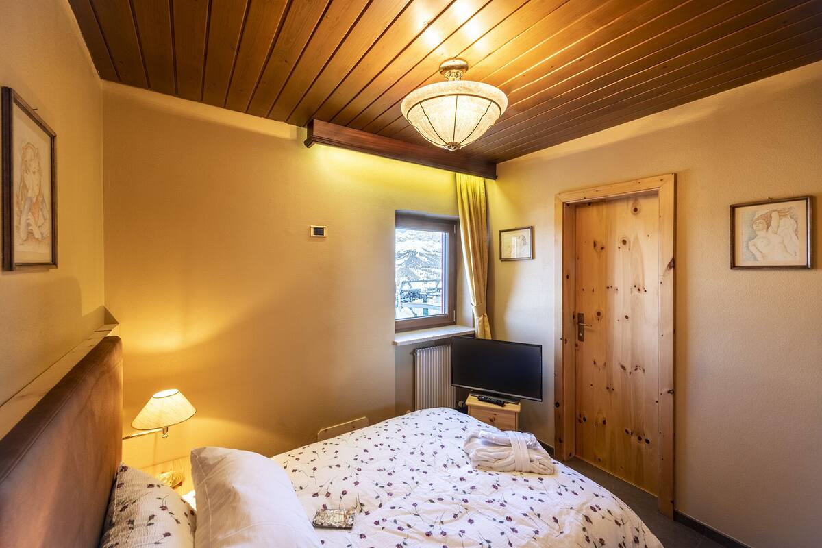 A hotel room with a wooden ceiling, a bed with floral bedding, a bedside lamp, a TV, a window with curtains, and a door.