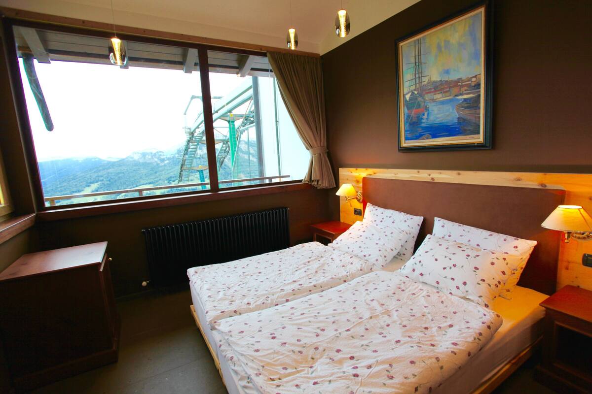 A hotel room with a large bed, two bedside tables, a window with a view of a bridge, and a painting on the wall.