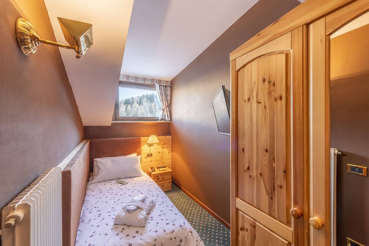 A small, cozy bedroom with a single bed, a nightstand, a lamp, a wooden wardrobe, and a window with a view of trees.