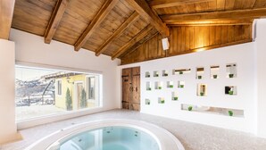 Une salle de spa dotée d’un bain à remous, de poutres en bois au plafond, d’une grande fenêtre offrant une vue sur un bâtiment et des montagnes, et d’un mur orné de plusieurs miroirs et de plantes vertes.