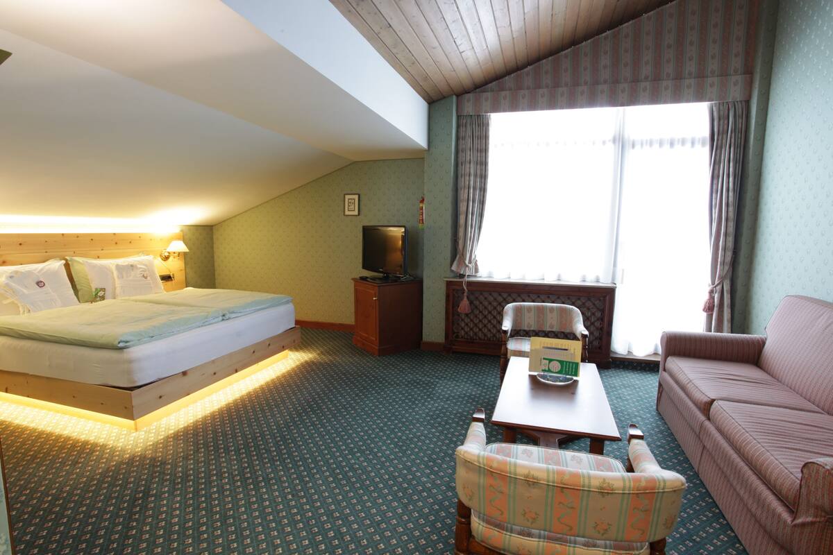 A hotel room with a bed, a sofa, a chair, a small table, a television, and a window with curtains.