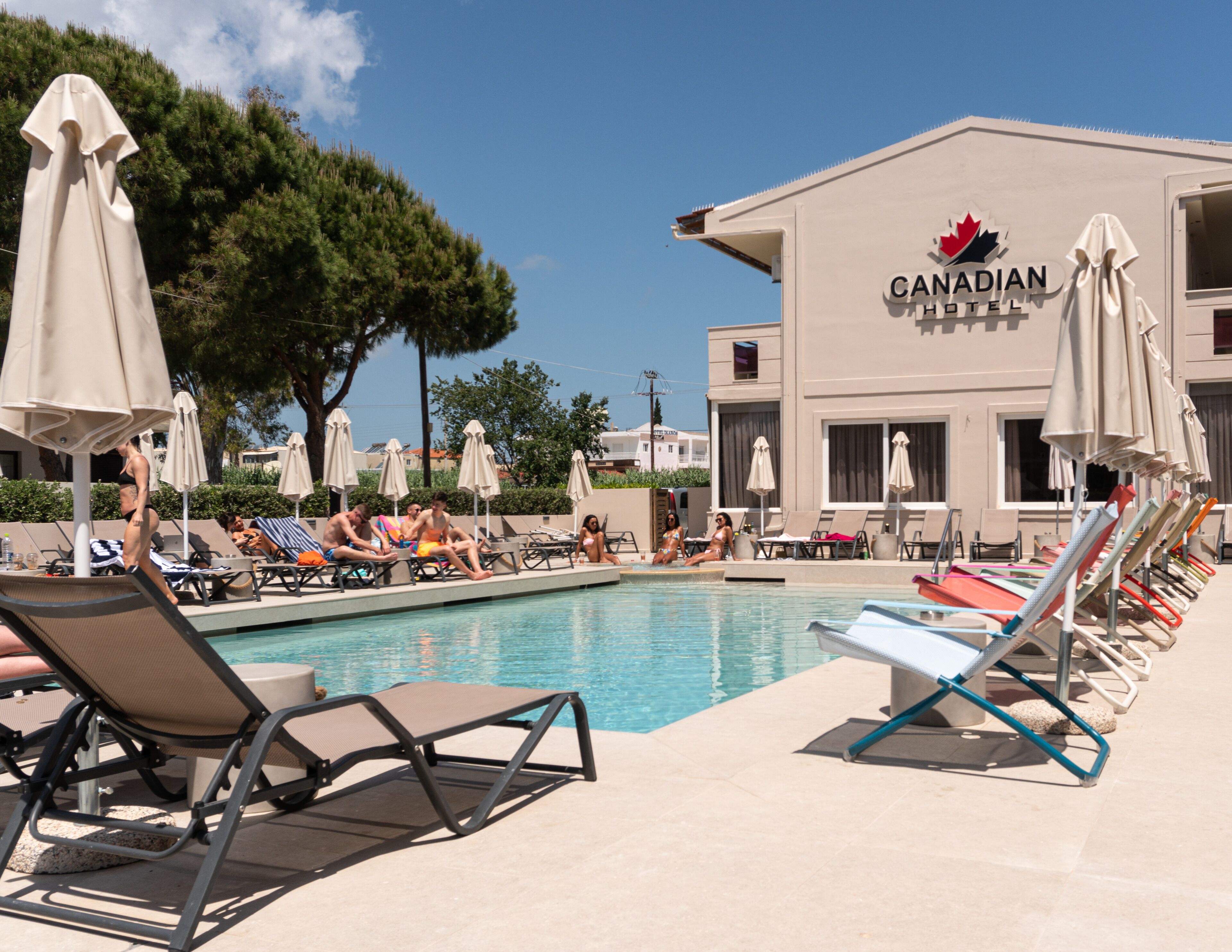 Et poolområde med liggestole og parasoller, en pool samt en bygning med det canadiske flag og hotelnavn.