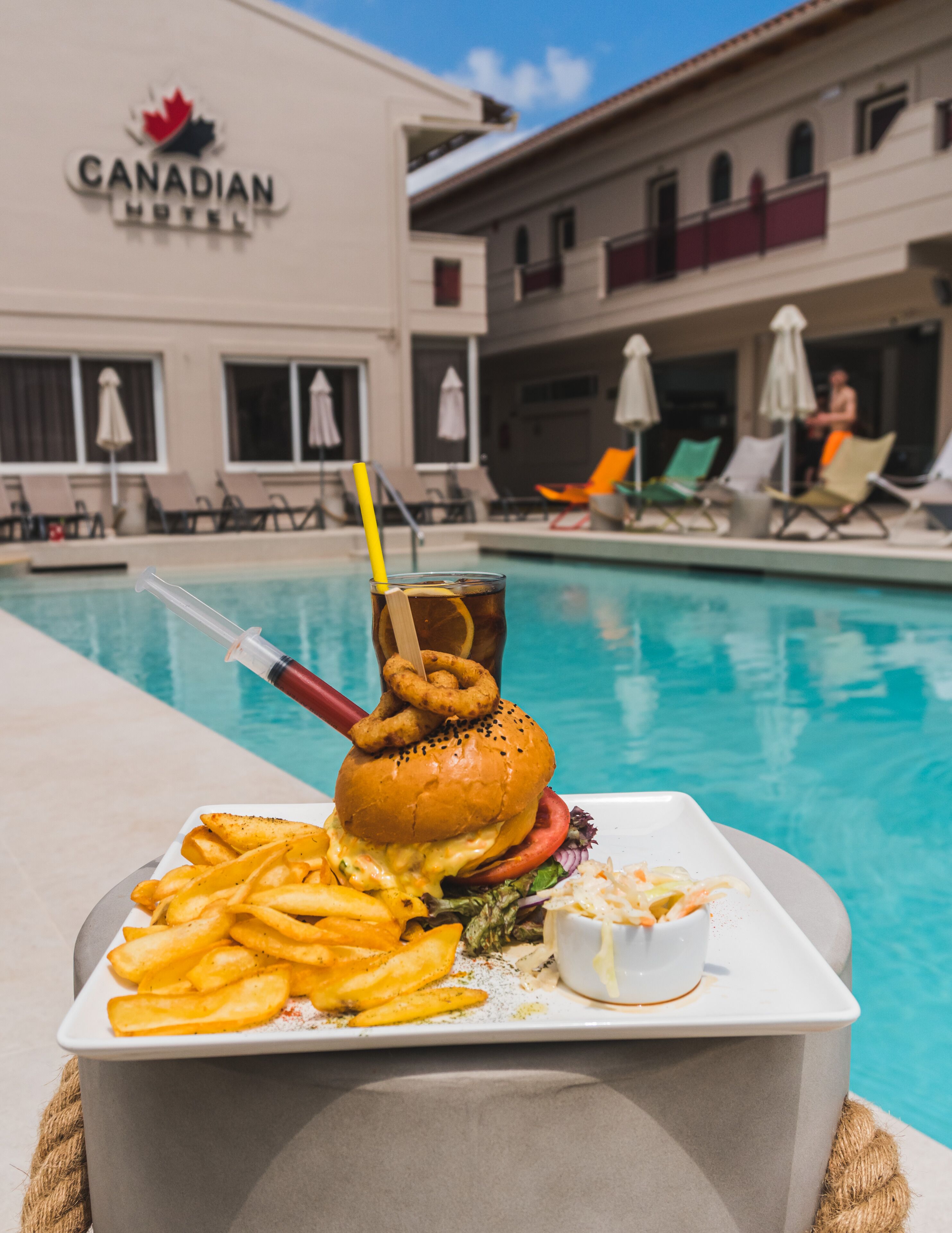 A dining table with a burger, fries, and a drink near a poolside area with lounge chairs and umbrellas.