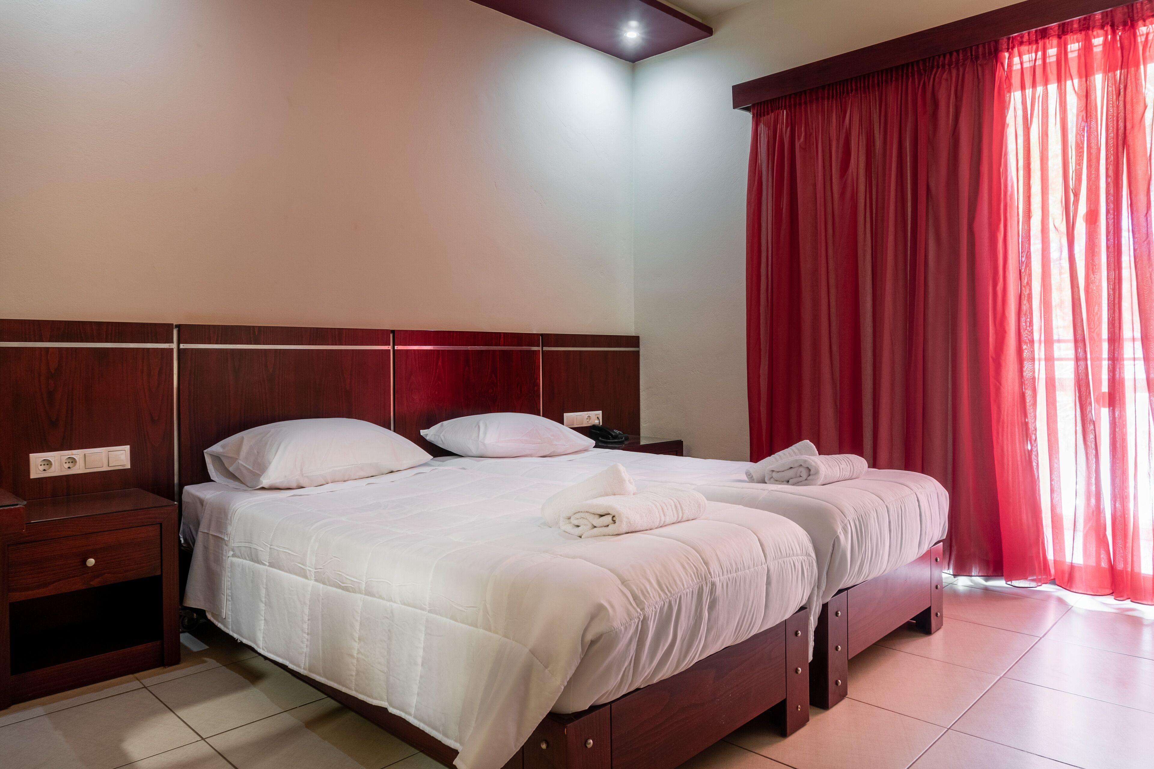 Habitación de hotel con dos camas, cabecero de madera, ropa de cama blanca, cortinas rojas y suelo de baldosas.