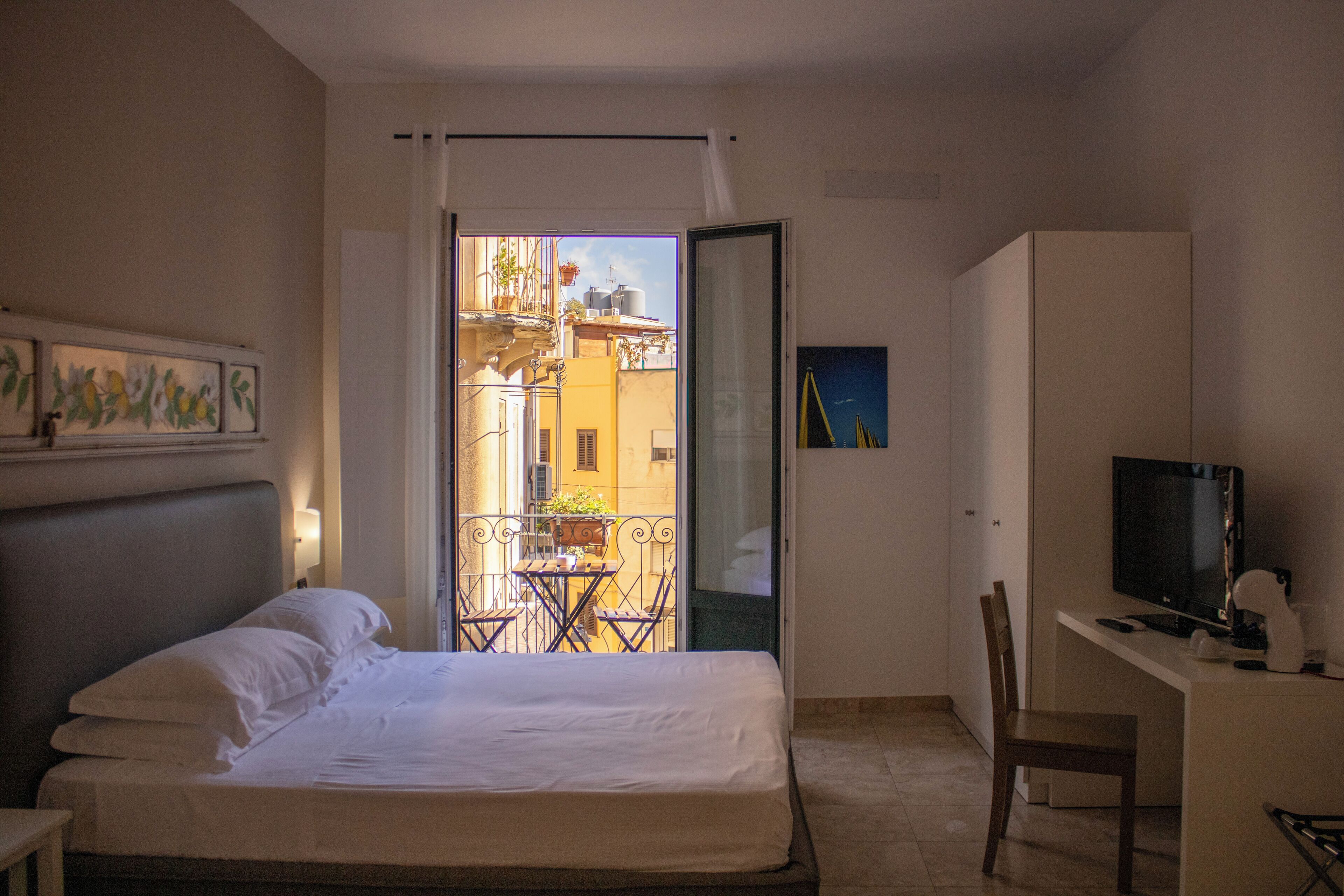 Habitación de hotel con cama, escritorio con televisor, una silla y balcón con vista a edificios.