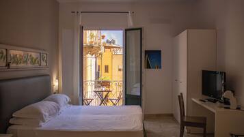 Habitación de hotel con cama, escritorio con televisor, una silla y balcón con vista a edificios.