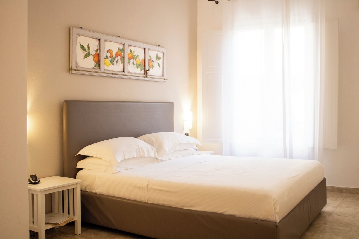 Un lit bien fait, avec une tête de lit grise, une petite table de chevet surmontée d’une lampe, et un tableau représentant des fruits accroché au mur.