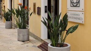 Un edificio con un letrero que dice "AL RESIDENCE SAN DOMENICO" y una bandera con diseño verde, blanco y rojo. Hay plantas en macetas frente a la entrada.