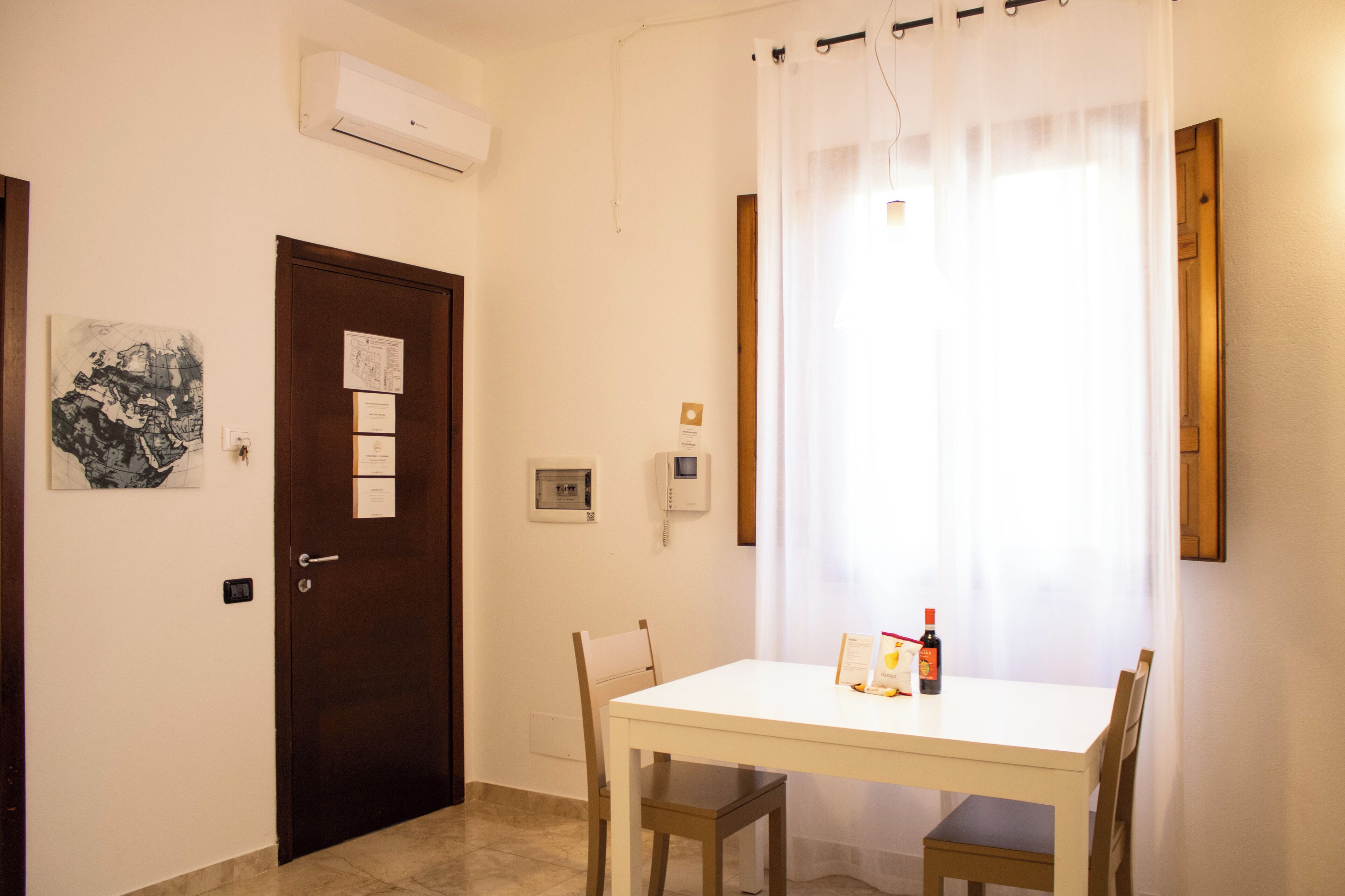 Uma pequena área de refeições com uma mesa e cadeiras brancas, uma porta com um mural de avisos e uma janela com cortinas leves.