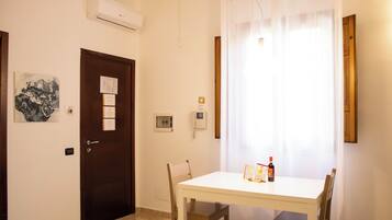 Uma pequena área de refeições com uma mesa e cadeiras brancas, uma porta com um mural de avisos e uma janela com cortinas leves.