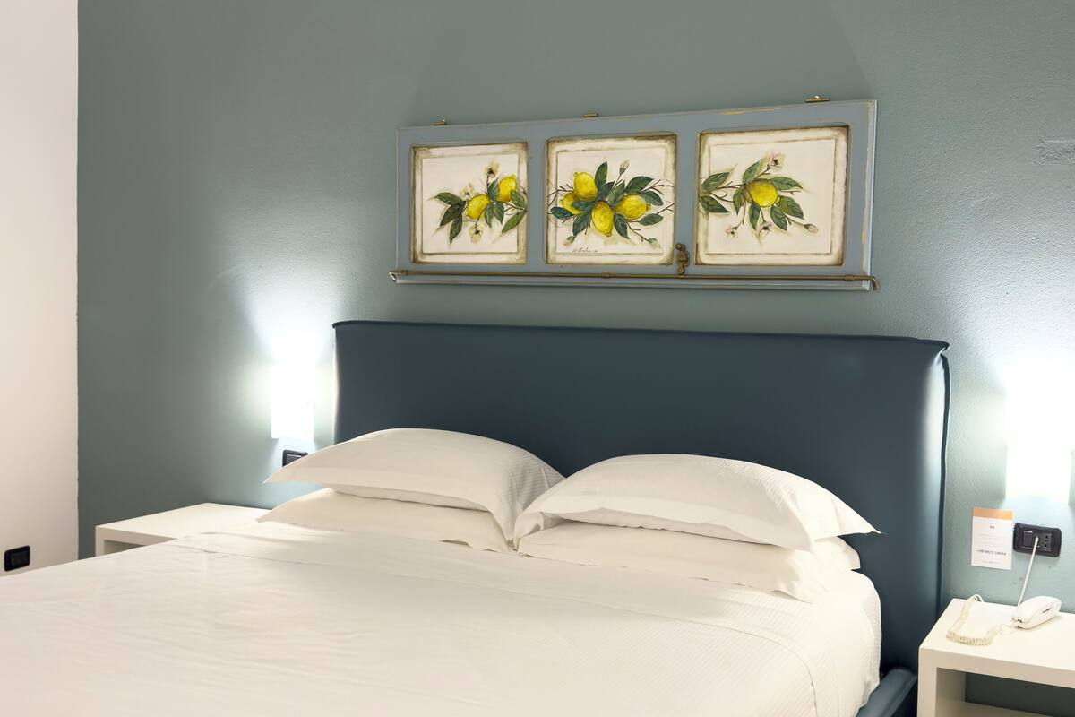 Una cama bien hecha con sábanas blancas, un cabecero oscuro y una mesita de noche con un teléfono y una tarjeta.