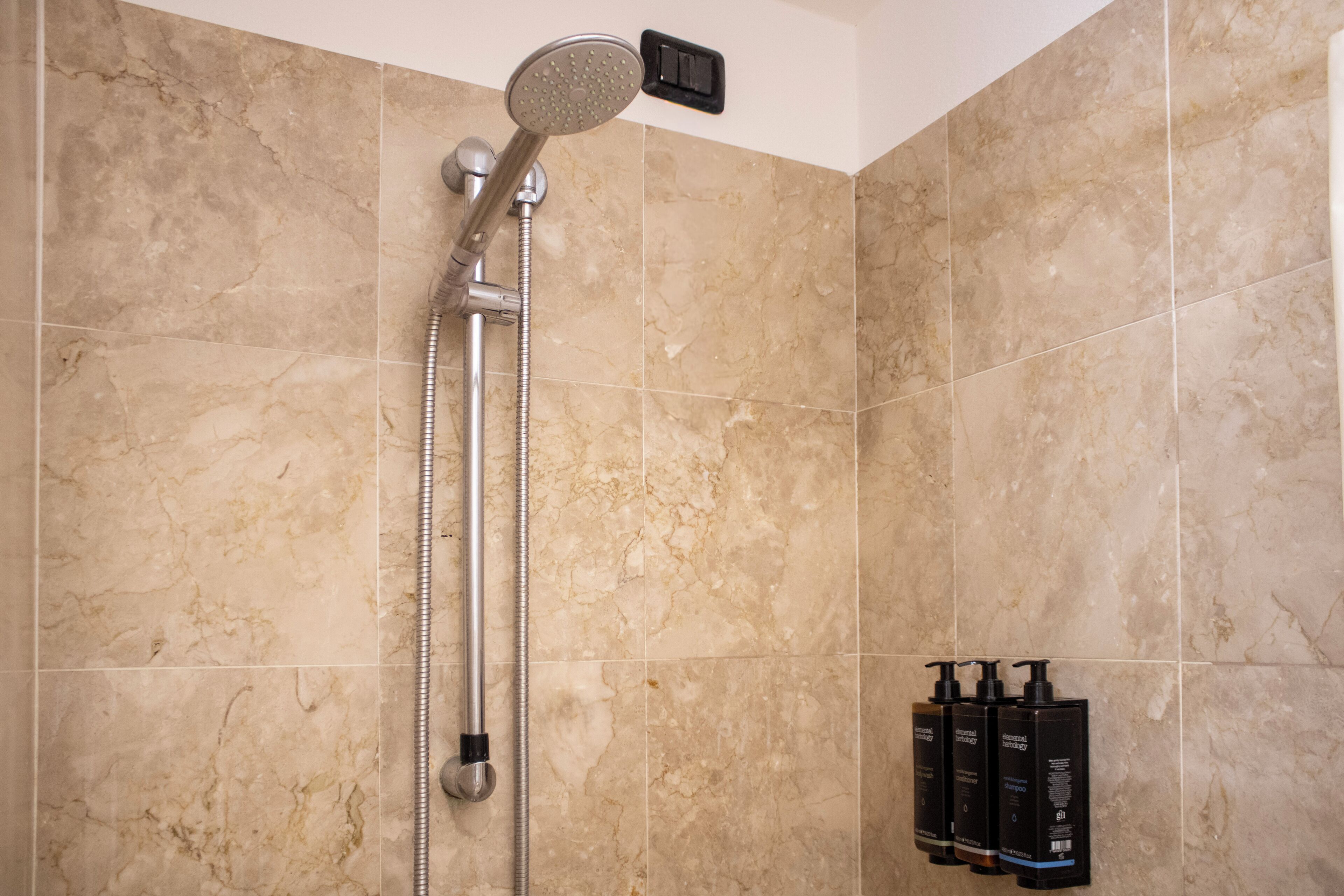 Ducha con cabezal de ducha manual y panel de control en la pared. Hay tres botellas de champú y acondicionador en la pared.