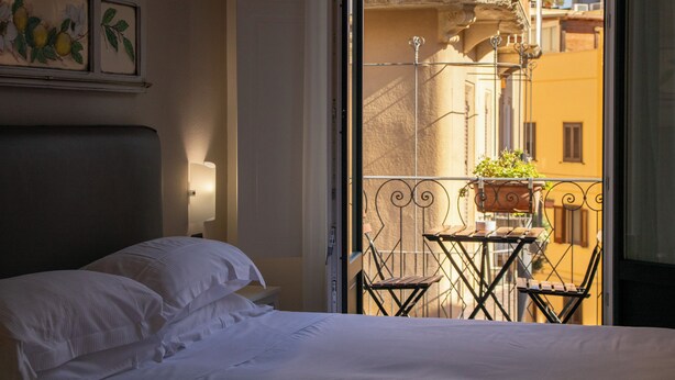 Habitación de hotel con cama, mesita de noche, lámpara de pared y vistas a un balcón con una pequeña mesa y sillas.