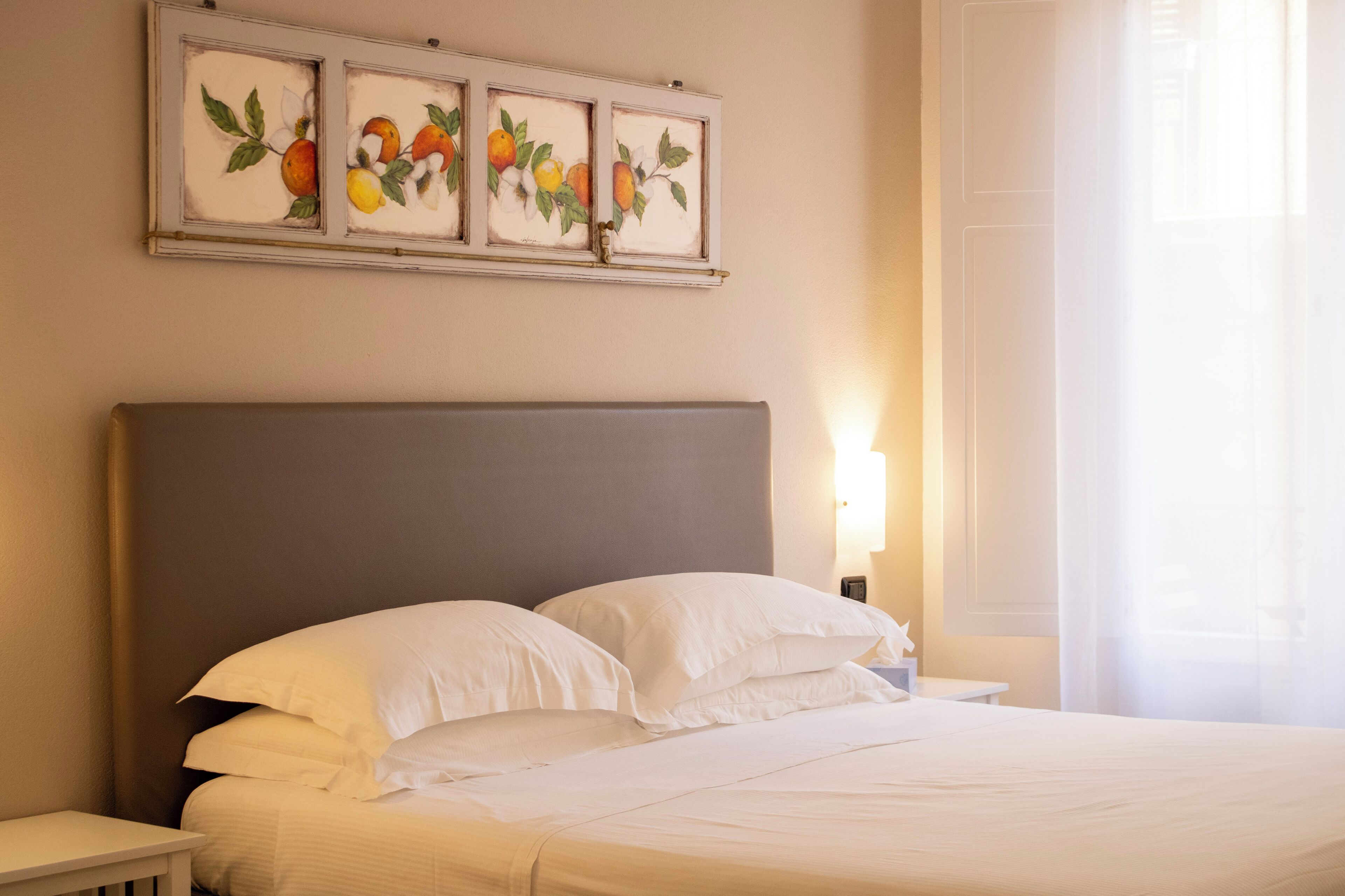 A neatly made bed with white linens, a grey headboard, and a bedside table with a lamp. Above the bed, there are three framed paintings of fruit.