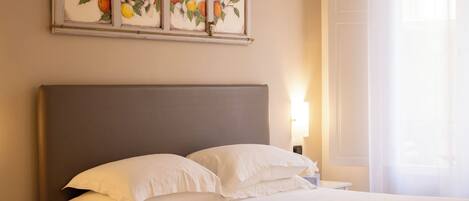 A neatly made bed with white linens, a grey headboard, and a bedside table with a lamp. Above the bed, there are three framed paintings of fruit.