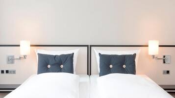 Habitación de hotel con dos camas, cada una con sábanas blancas y almohada azul oscuro. Hay lámparas de pared a cada lado de las camas.