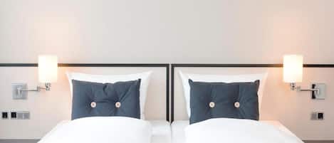 A hotel room with two beds, each with white linens and a dark blue pillow. There are wall-mounted light fixtures on either side of the beds.